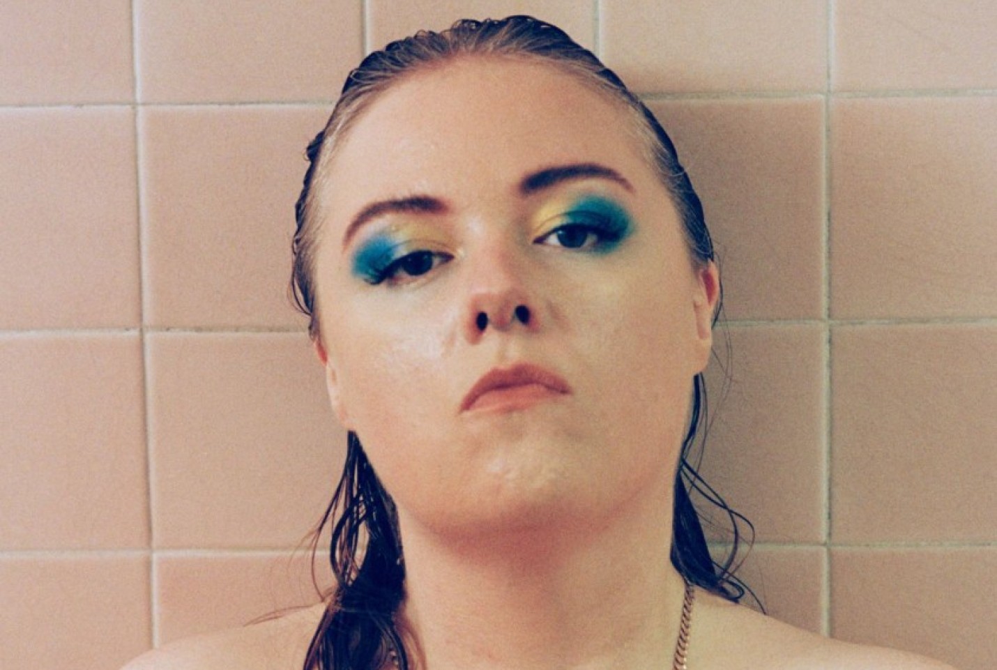 Emma-Jean Thackray sitting in front of a tiled wall with wet hair and blue and yellow eyeshadow.